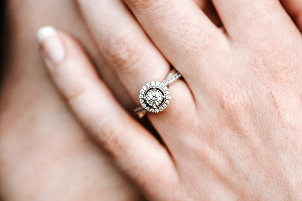 A close-up of a woman’s finger, adorned with a halo style engagement ring.