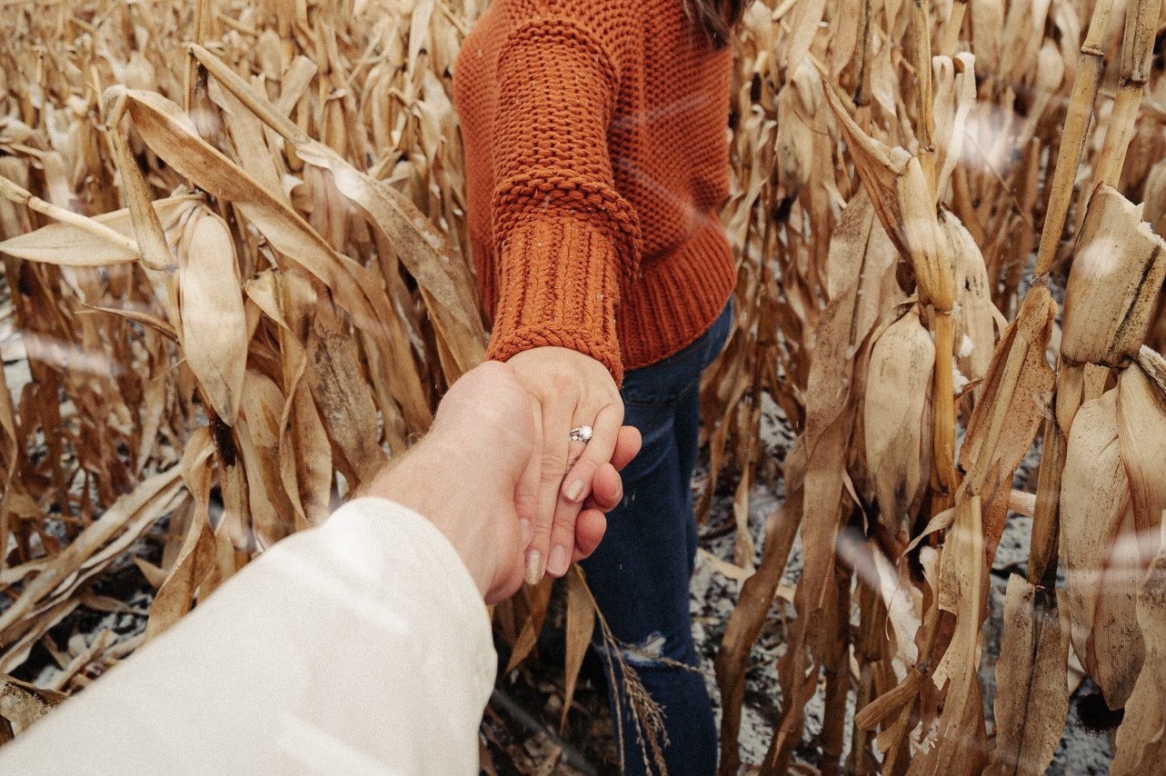 A woman playfully leading her partner into a cornfield, an elegant engagement ring on her finger.