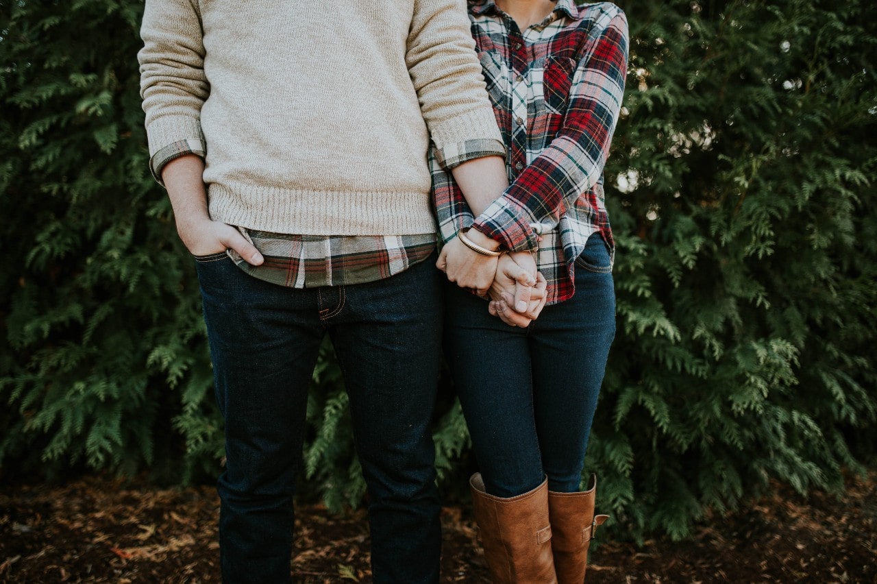 A close-up of a warmly dressed couple holding hands in a wooded area, a cuff bracelet on her wrist.