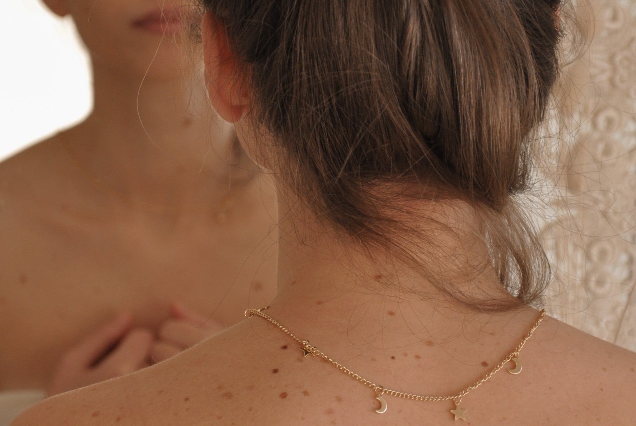 A posterior view of a woman looking in the mirror, wearing a gold necklace with star and moon motifs.