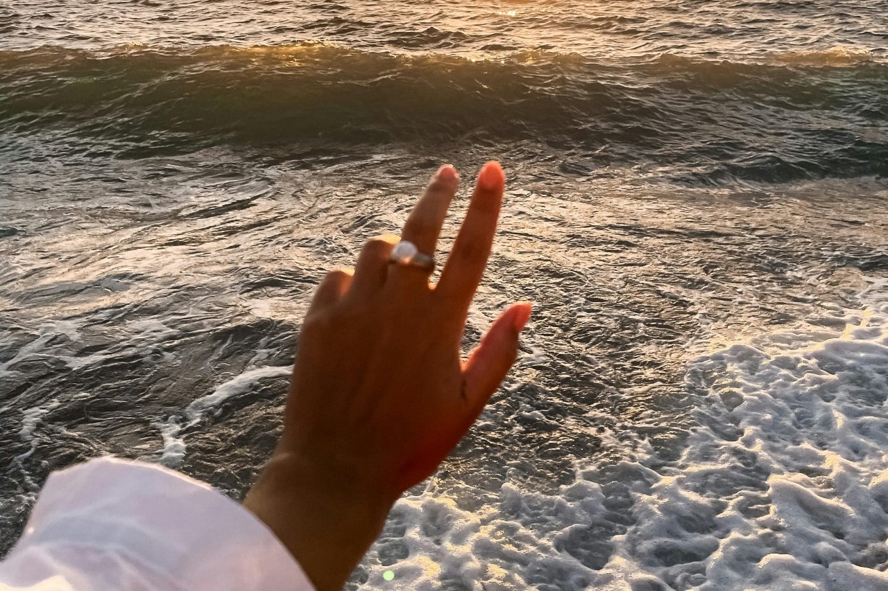 A woman’s hand adorned with a pearl fashion ring, extended towards the ocean.