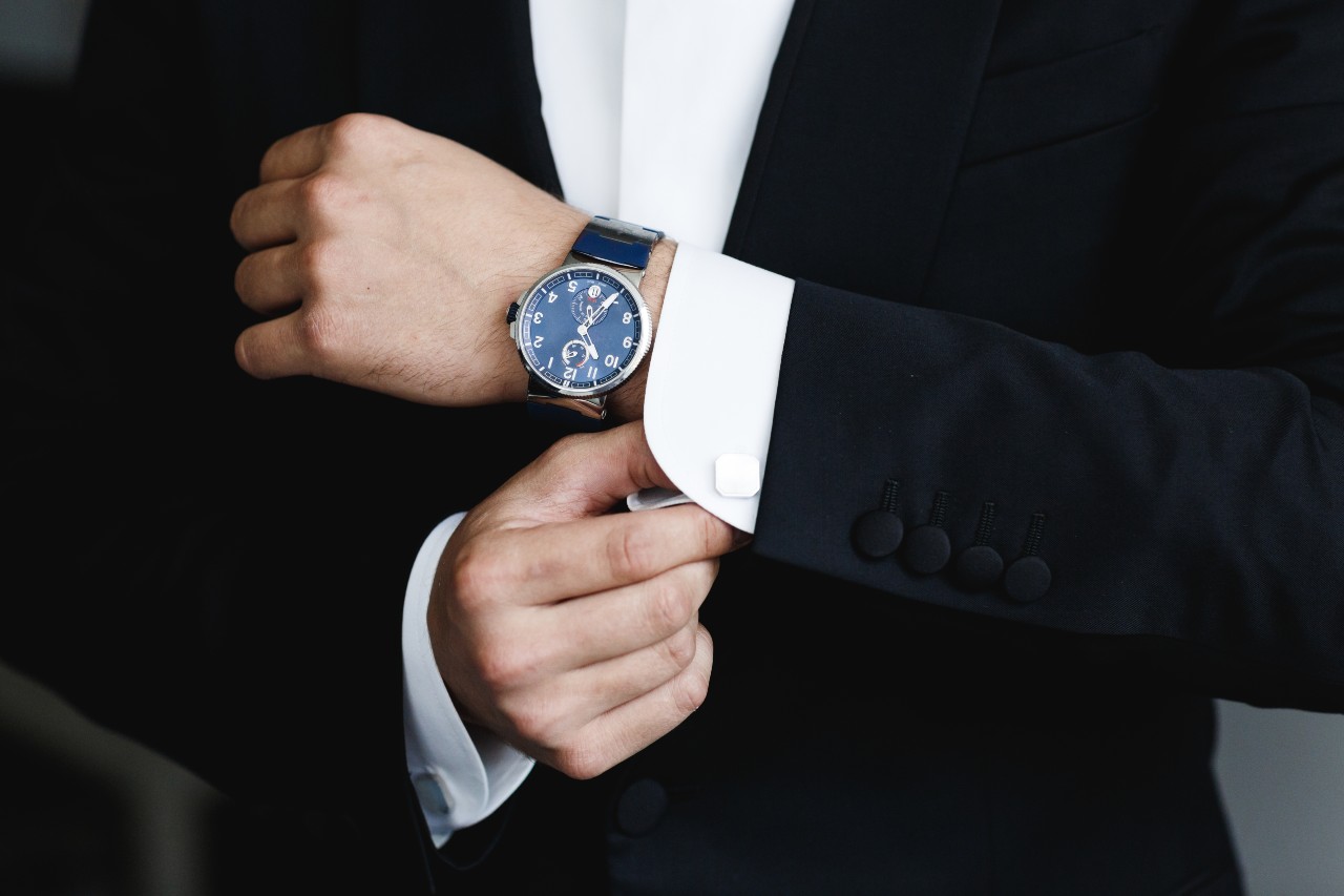 A man in a black suit adjusts his white shirt cuff, showcasing a wristwatch with blue dial.