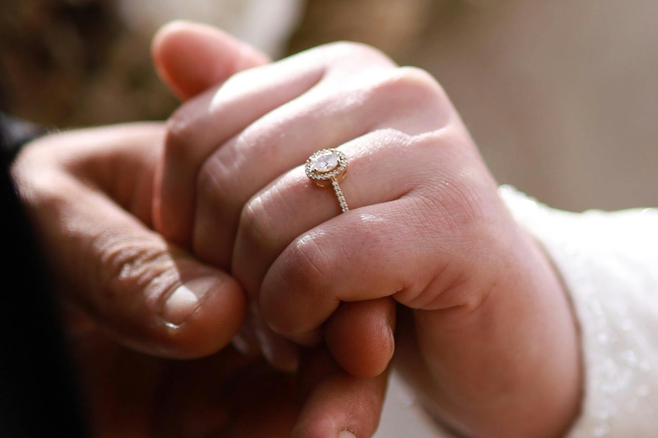 a man’s hand holding a woman’s that is adorned with an oval cut halo set engagement ring