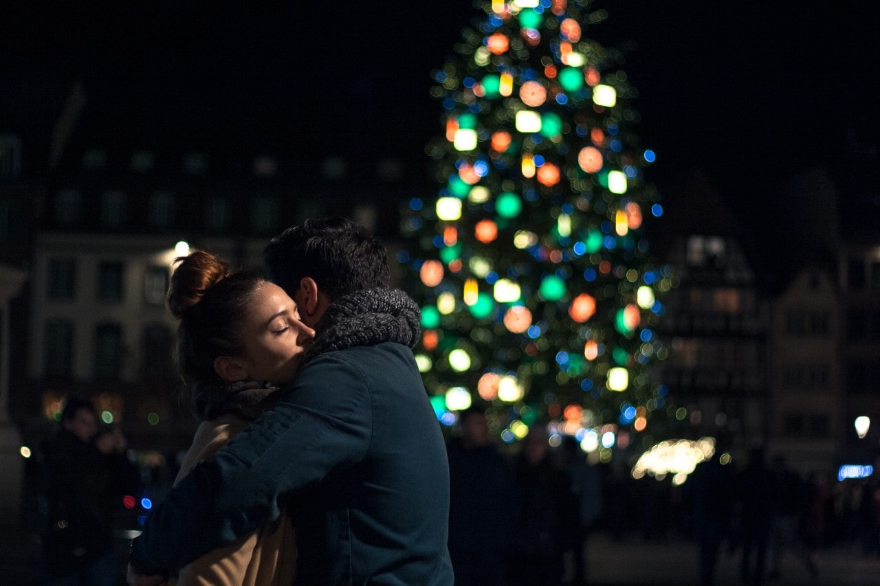 A recently engaged couple embracing before a well-lit Christmas tree.