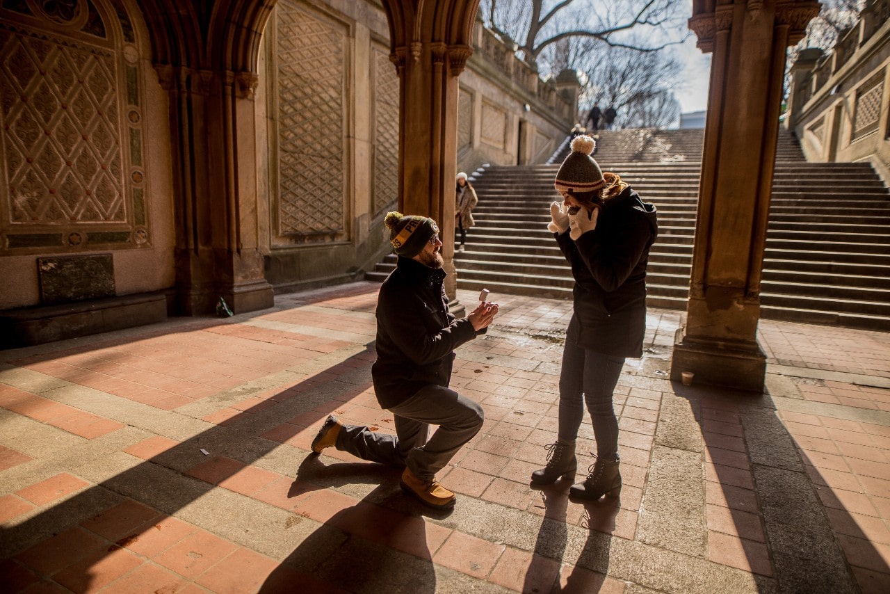 A man proposing to his partner in a scenic walkway, both warmly dressed for winter.