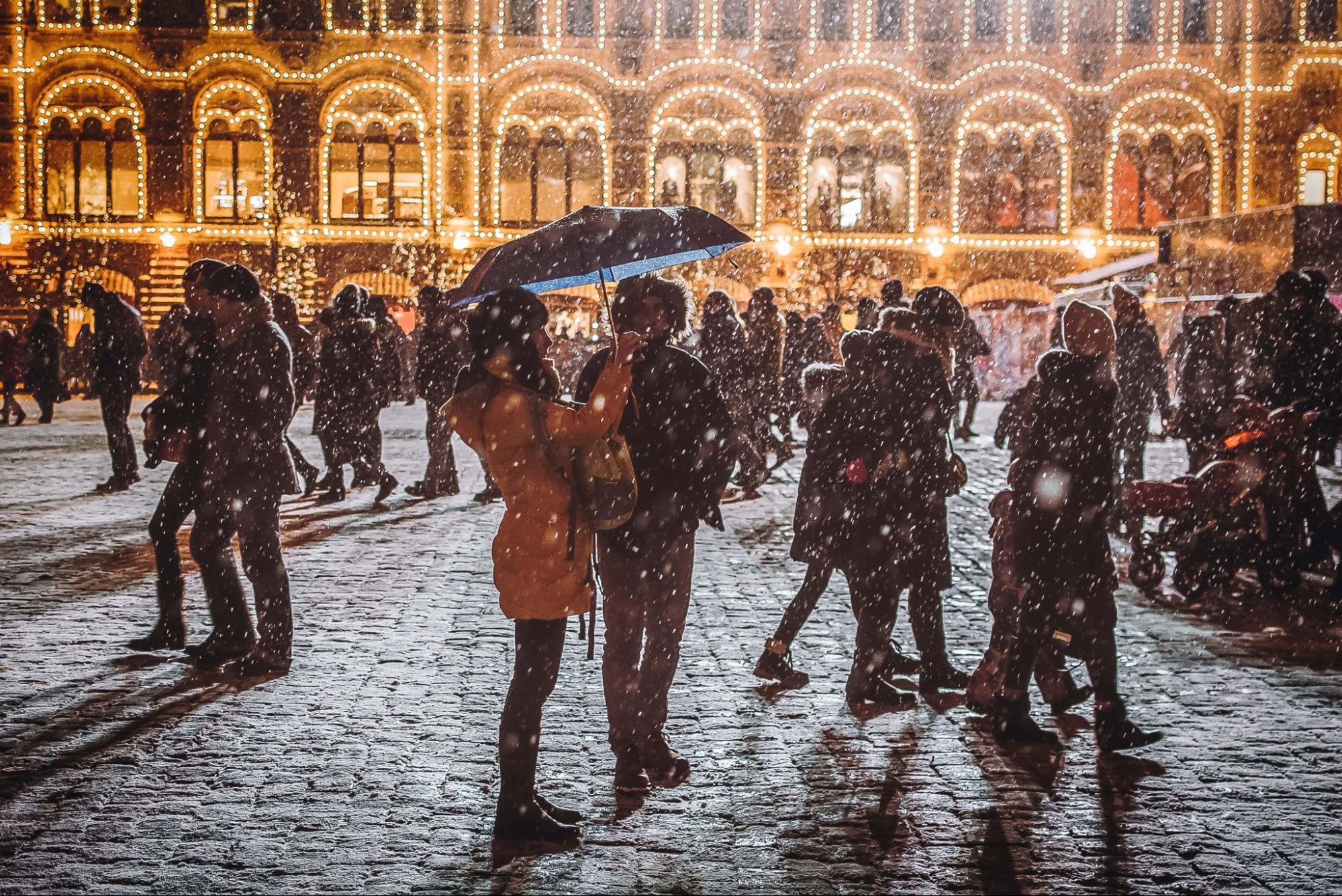 A wide shot of a warmly dressed couple among a crowd in front of a festively lit building as snow falls.