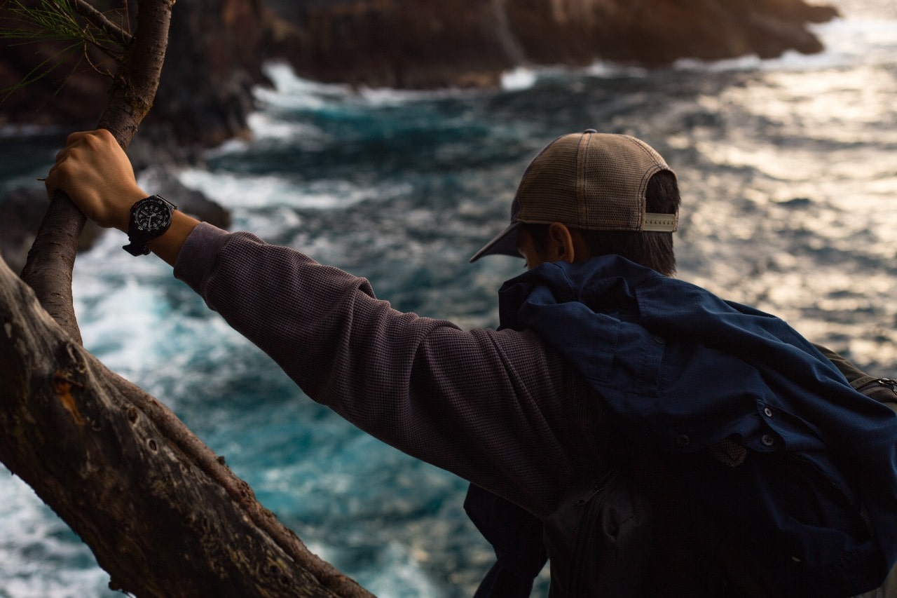 A hiker watches a river as he holds onto a tree branch.