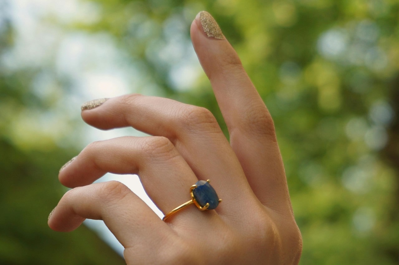 a woman’s hand raised to the sky, adorned with a yellow gold solitaire engagement ring with a blue stone