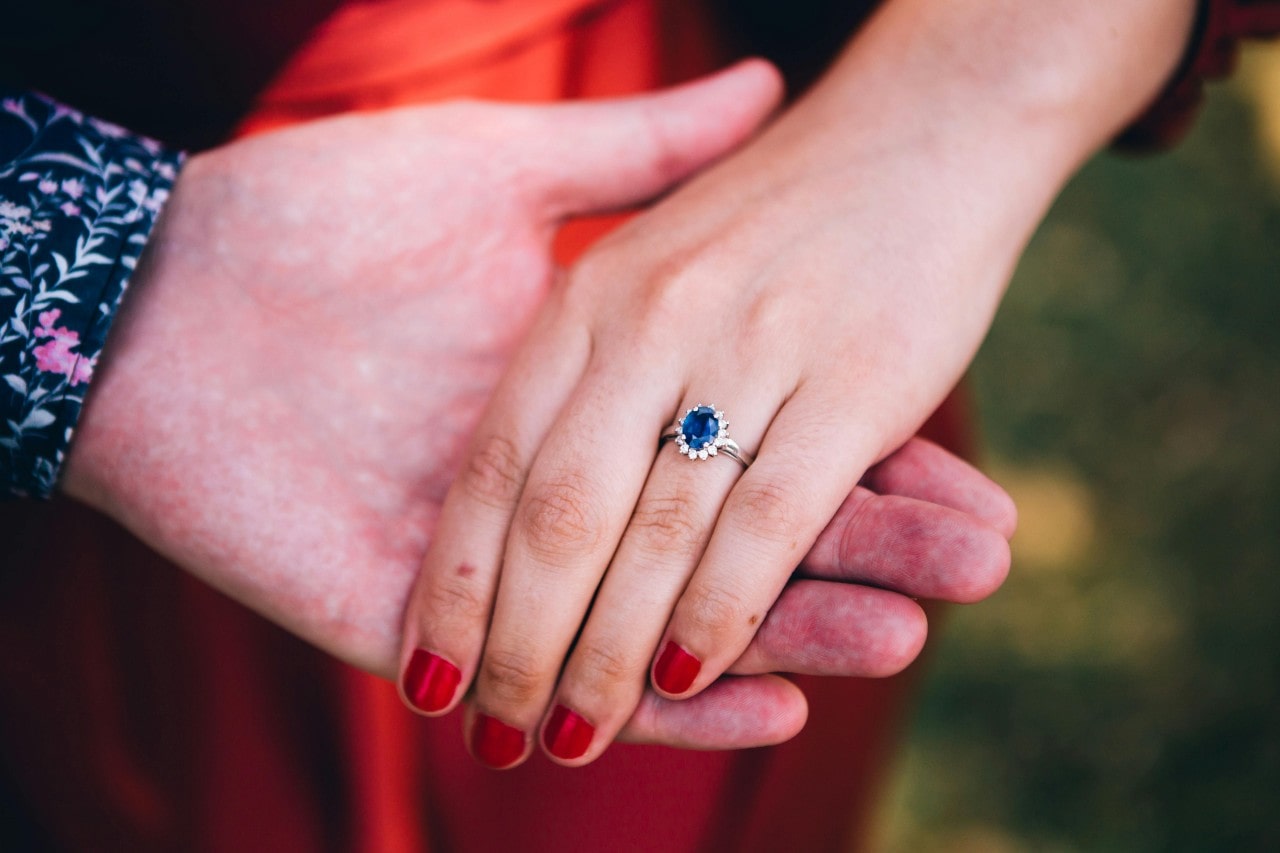 A man’s hand holding a woman’s hand, adorned with a sapphire, halo set engagement ring.