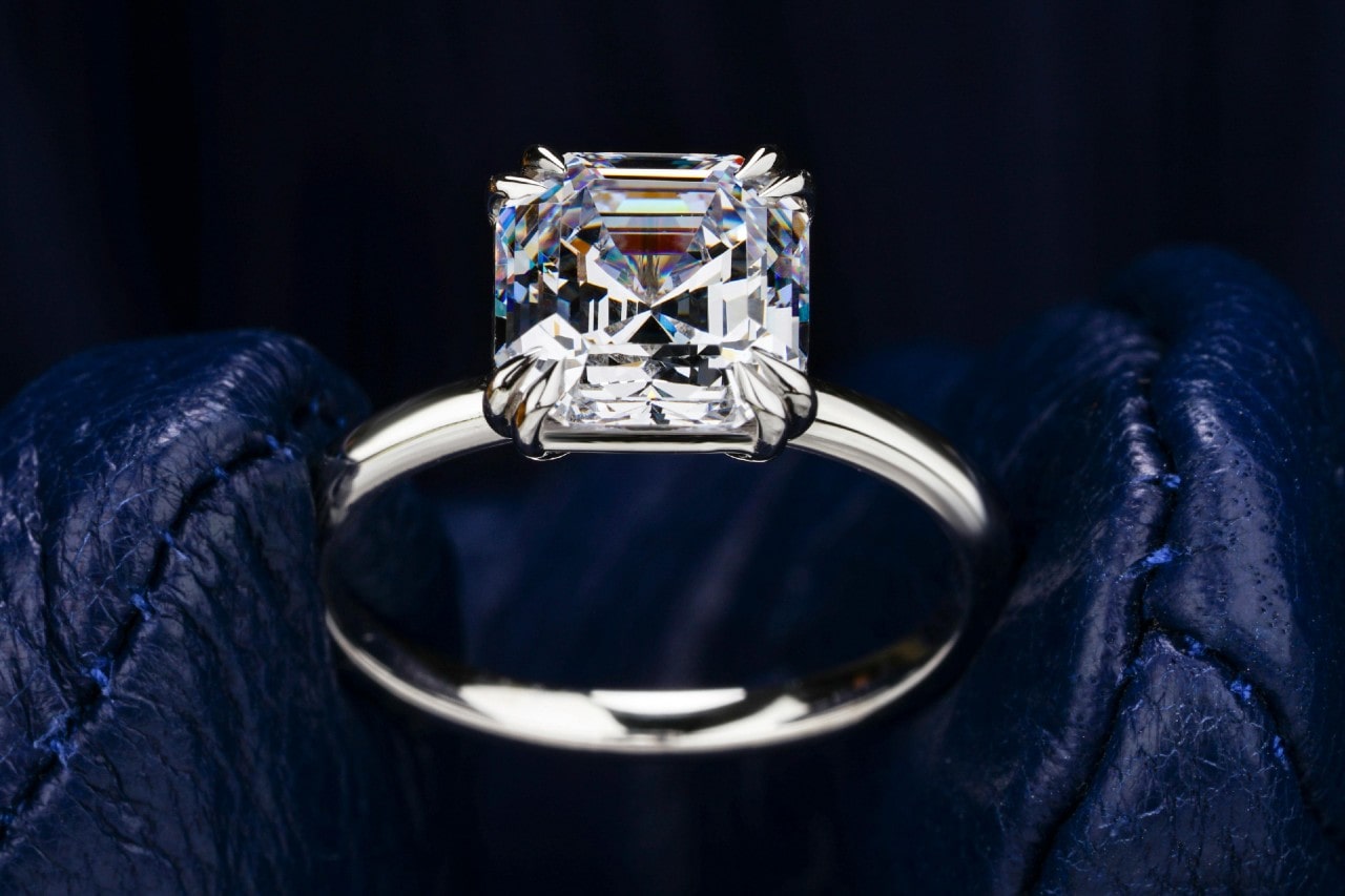 A close-up of an asscher cut solitaire engagement ring on a dark leather backdrop.